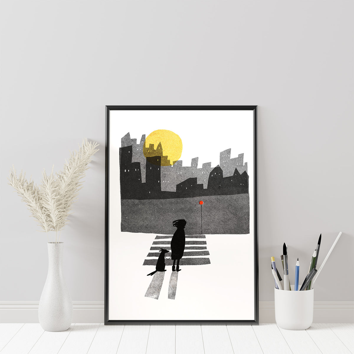 Across art print by Magda Brol: a woman and her dog crossing a zebra crossing at night under a yellow moon, with a dark city skyline, in a black frame beside a vase of dried grass and a pot of brushes