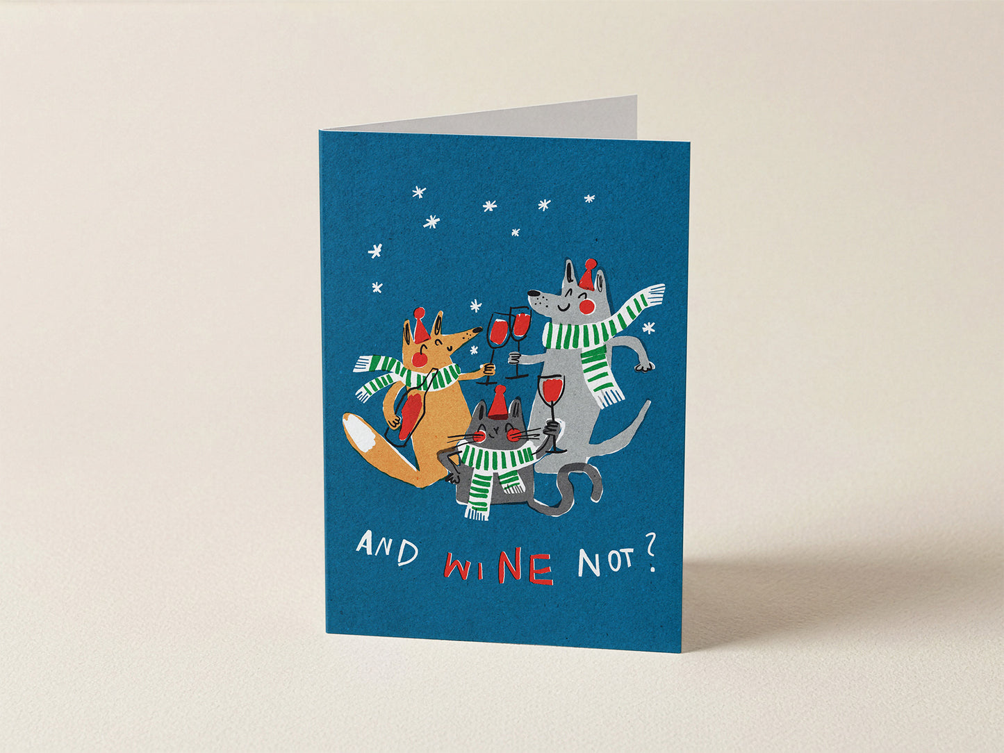 A blue greeting card with a whimsical illustration of three dogs wearing green-and-white striped scarves and red hats, each holding a wine glass, with the text 'AND WINE Not?' on a light beige background.