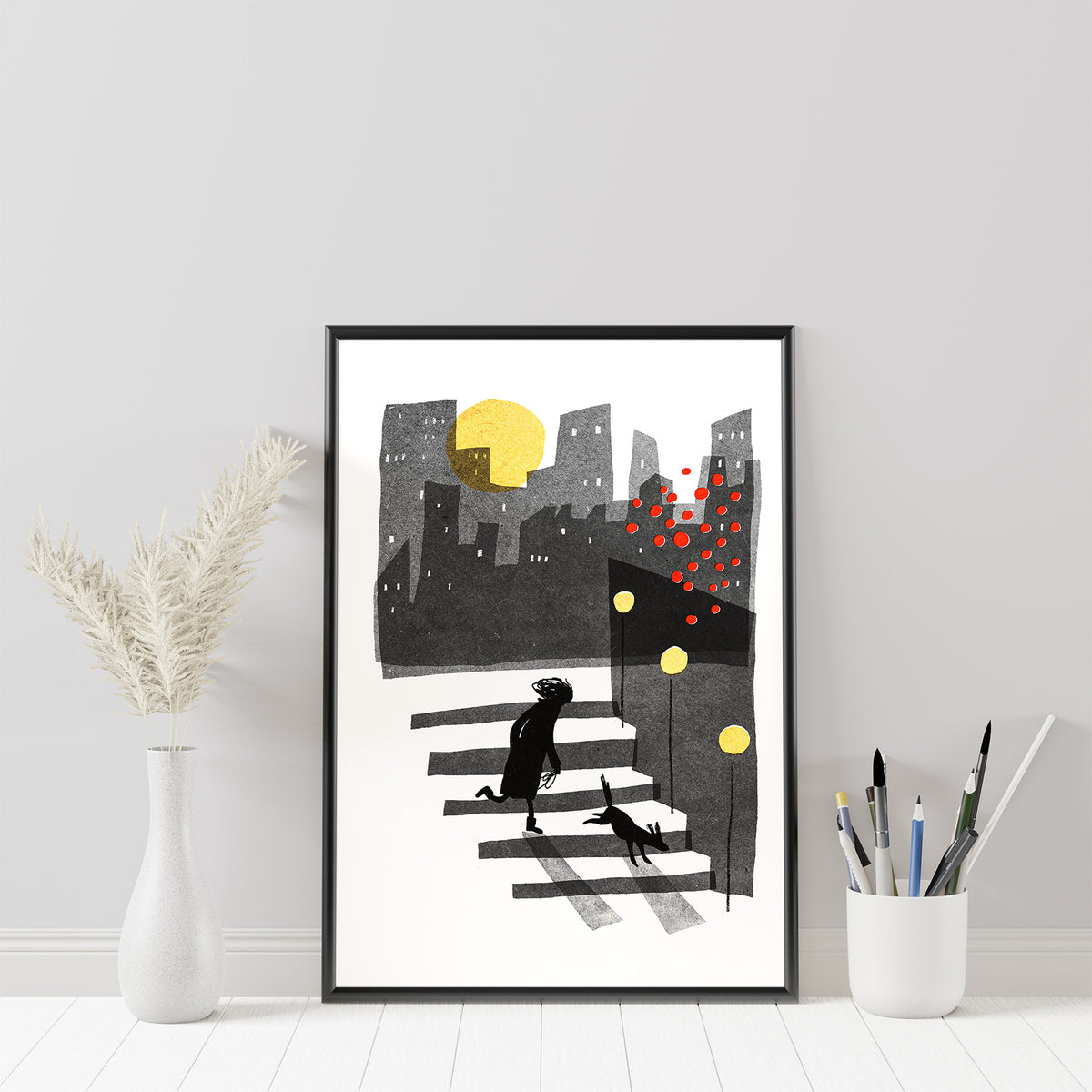 Down art print by Magda Brol: a woman and her dog walking down steps at night past red and yellow lights, under a yellow moon and city skyline, in a black frame beside a vase of dried grass and a pot of brushes